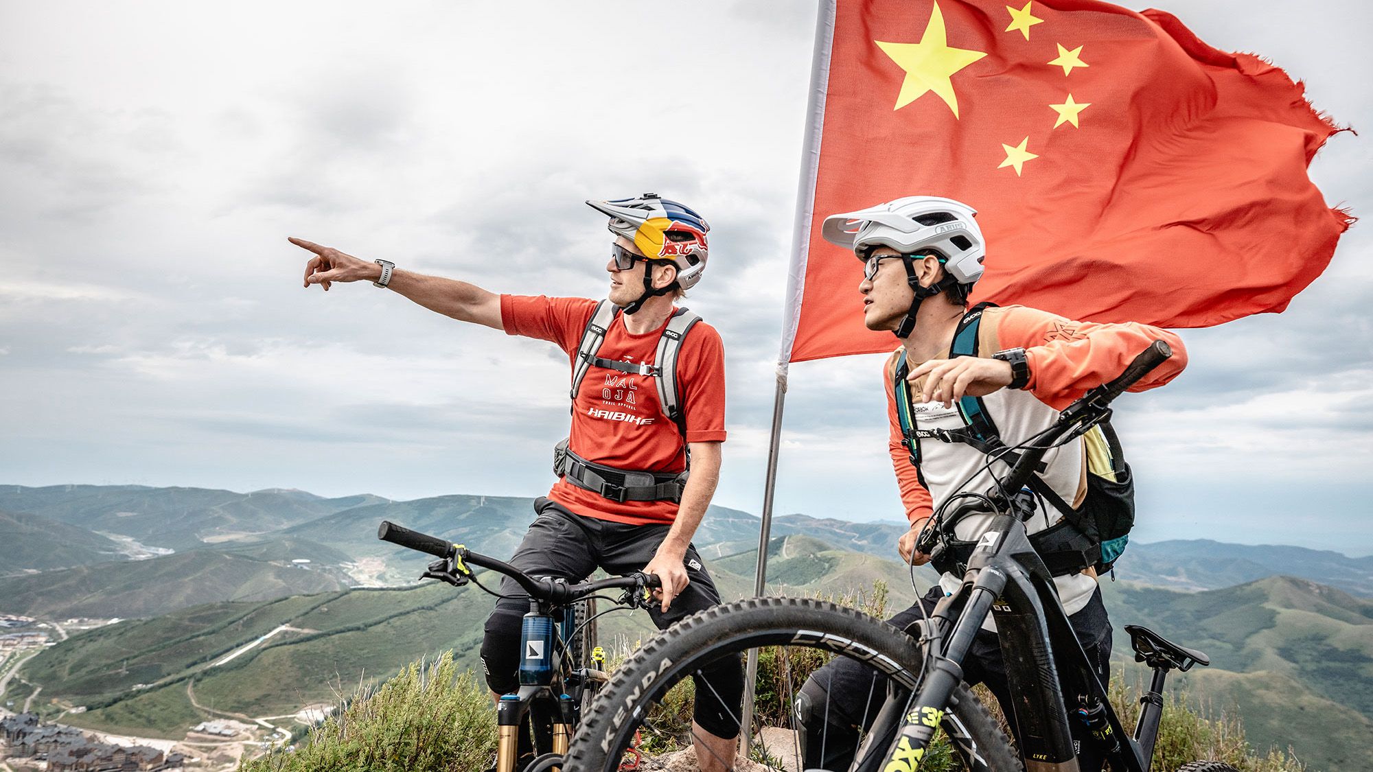 Zwei Mountainbiker auf einem Berggipfel mit der chinesischen Fahne im Hintergrund.