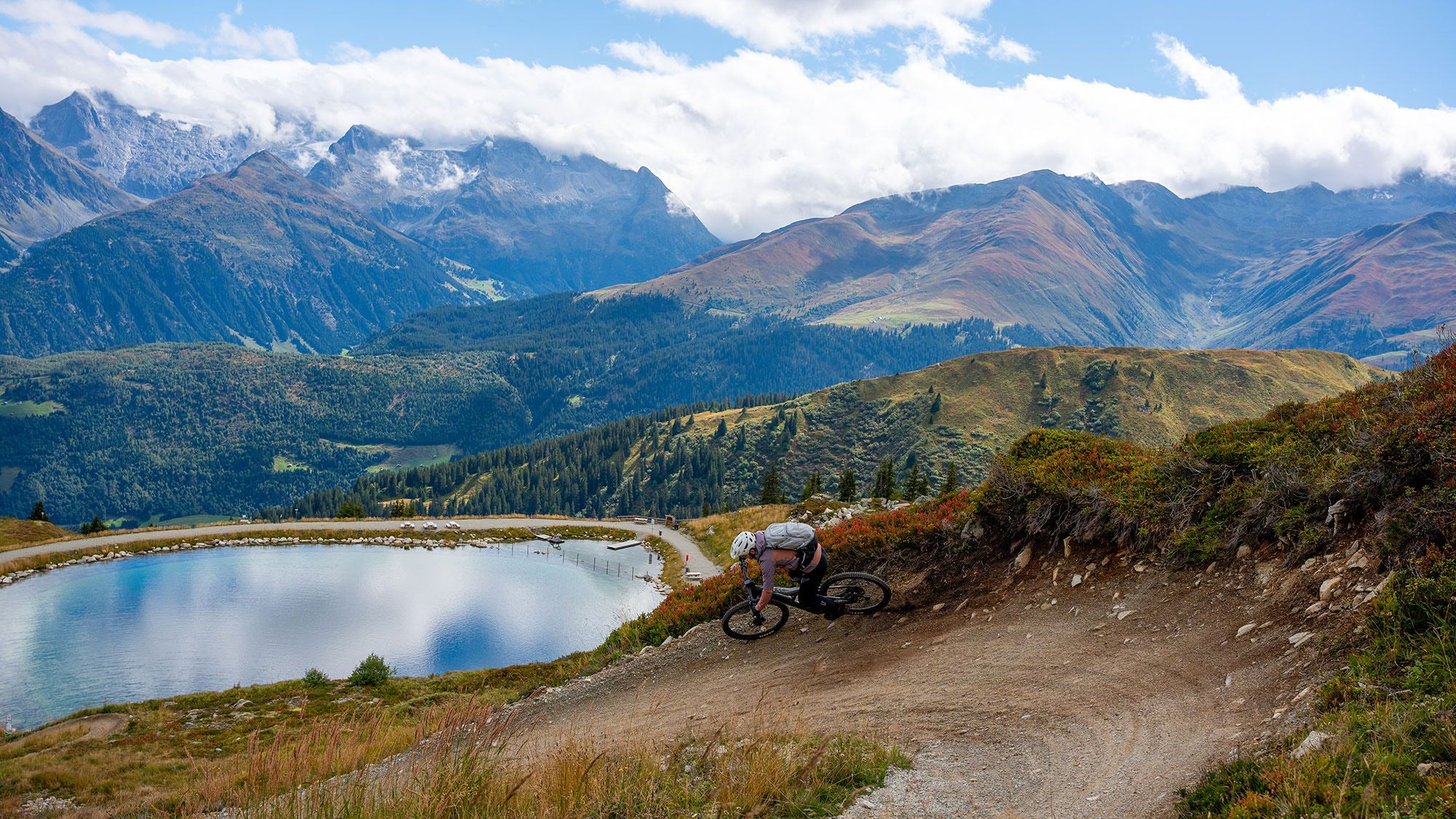 Ein Mountainbiker in einer Kurven, dahinter ein See mit Bergpanorama
