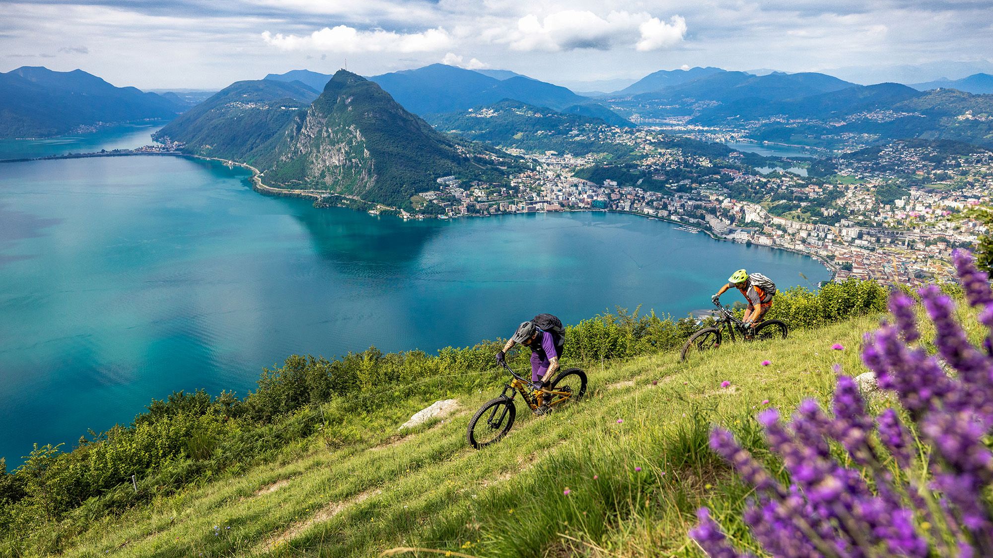 Zwei Mountainbiker fahren einen Trail mit Blick auf einen See.